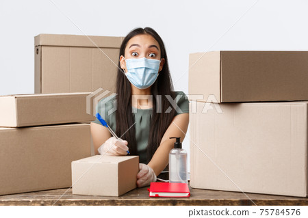 Small business owners, e-commerce and covid-19 preventing virus concept. Surprised asian businesswoman in medical mask and gloves looking astonished while writing shipping info on order box Small business owners, e-commerce and covid-19 preventing virus concept. Surprised asian businesswoman in medical mask and gloves looking astonished while writing shipping info on order box 75784576