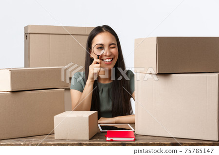 Small business owners, startup and e-commerce concept. Smiling asian businesswoman processing client orders, selling online, checking parcels with magnifying glass, ensuring best quality Small business owners, startup and e-commerce concept. Smiling asian businesswoman processing client orders, selling online, checking parcels with magnifying glass, ensuring best quality 75785147