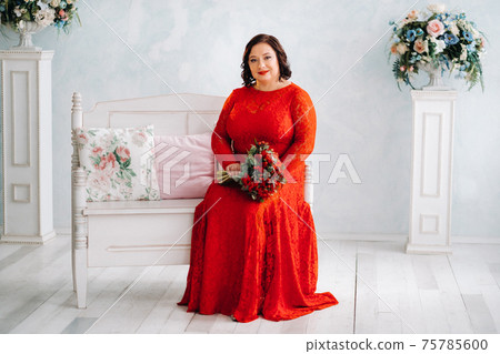 a woman in a red dress sits on a sofa and holds a bouquet of red roses and strawberries in the interior. a woman in a red dress sits on a sofa and holds a bouquet of red roses and strawberries in the interior. 75785600