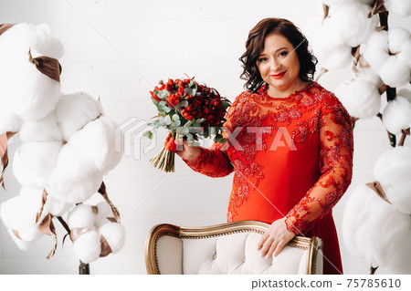 a woman in a red dress stands and holds a bouquet of red roses and strawberries in the interior. 75785610