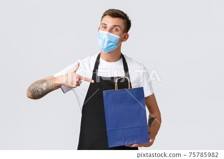 Retail store, shopping during covid-19 and social distancing concept. Friendly handsome salesman in black apron and medical mask pointing finger at eco-bag, white background 75785982