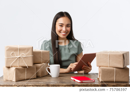 Small business owners, startup and e-commerce concept. Smiling asian businesswoman work from home, packing sold items, wrap boxes with orders for shipping, manage online store via digital tablet Small business owners, startup and e-commerce concept. Smiling asian businesswoman work from home, packing sold items, wrap boxes with orders for shipping, manage online store via digital tablet 75786397