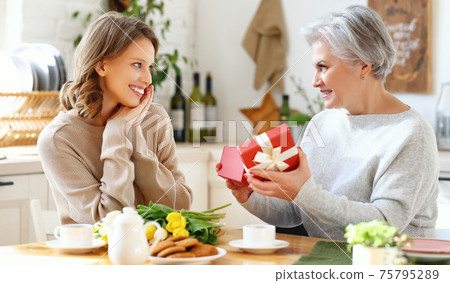 Positive aged woman giving present to smiling daughter during breakfast at home Positive aged woman giving present to smiling daughter during breakfast at home 75795289