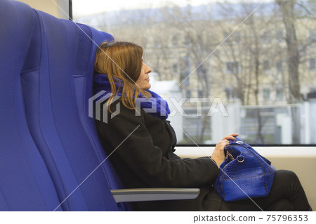 A young brunette woman sits and looks out the train window at the cityscapes. Holding a blue handbag. A blue scarf and blue seats. 75796353