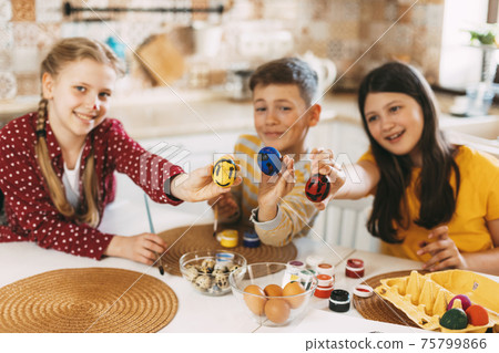 Funny and cheerful children sitting at the table paint Easter eggs in different colors for Easter Funny and cheerful children sitting at the table paint Easter eggs in different colors for Easter 75799866