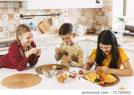 Two sisters and a brother are sitting at the table and painting Easter eggs in different colors for Easter, laughing and indulging Two sisters and a brother are sitting at the table and painting Easter eggs in different colors for Easter, laughing and indulging 75799892