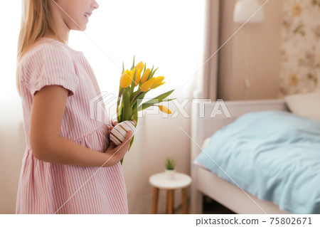 Easter concept. Girl in bunny ears and pink dress holding yellow tulips and egg 75802671