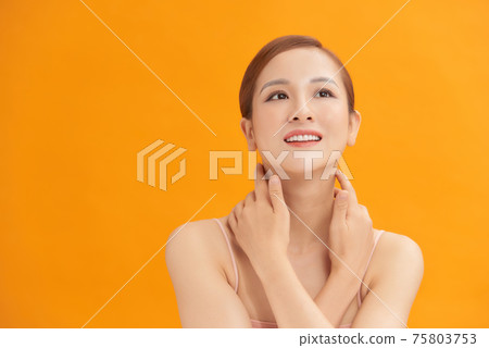 Charming woman touching her perfect soft face skin with fingers, smiling at camera over yellow background 75803753