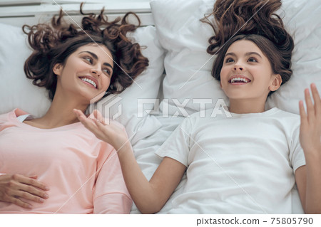 Daughter cheerful gesturing and interested mom lying on bed Daughter cheerful gesturing and interested mom lying on bed 75805790