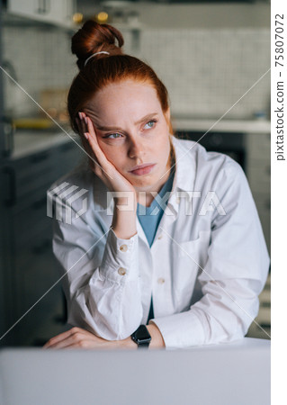 Close-up portrait of tired young redhead woman sitting at desk in cozy living room. 75807072