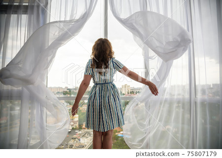 Rear view of a young joyful woman wearing fashion dress and holding the curtains open to look out of large light window at home, turning to look and smile at camera, interior 75807179
