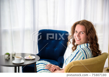 Young woman at home sitting on modern chair in front of window relaxing in her living room and drinking coffee or tea with sweets or desert. Relax before new business day 75807180