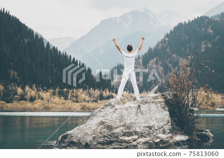 A man stands on a large stone and raises his hands to the sky. Snowy peaks of the mountains. , Issyk city, Kazakhstan A man stands on a large stone and raises his hands to the sky. Snowy peaks of the mountains. , Issyk city, Kazakhstan 75813060