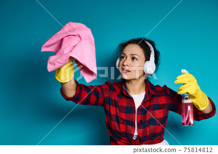 Woman in headphones and yellow rubber gloves holds detergent spray and cleaning rag, simulating cleaning surface. Woman in headphones and yellow rubber gloves holds detergent spray and cleaning rag, simulating cleaning surface. 75814812