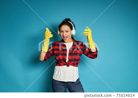 Young african woman in red plaid shirt with headphones on her head sings, posing on a blue background with cleaning sponges in her hands 75814814