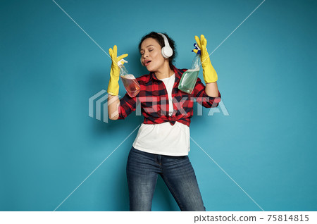 Young african woman wearing red plaid shirt and blue jeans with headphones on her head sings, posing on a blue background with cleaning sponges in her hands Young african woman wearing red plaid shirt and blue jeans with headphones on her head sings, posing on a blue background with cleaning sponges in her hands 75814815