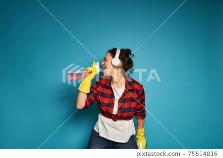 African American woman, housewife, with headphones holding cleaning sprays in her hands and singing with her eyes closed, enjoying her homework. African American woman, housewife, with headphones holding cleaning sprays in her hands and singing with her eyes closed, enjoying her homework. 75814816