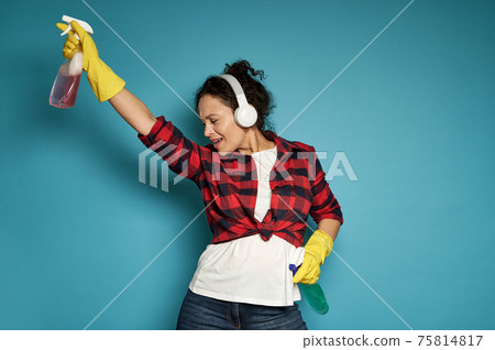 Hispanic woman, housewife, with headphones holding cleaning sprays in her hands and dancing with her eyes closed, enjoying her homework. 75814817