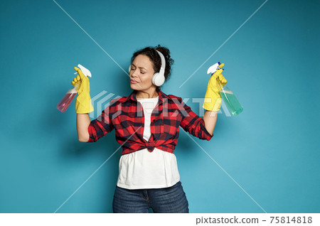 Hispanic woman, housewife, with headphones holding cleaning sprays in her hands and listening to music with her eyes closed, enjoying her homework Hispanic woman, housewife, with headphones holding cleaning sprays in her hands and listening to music with her eyes closed, enjoying her homework 75814818