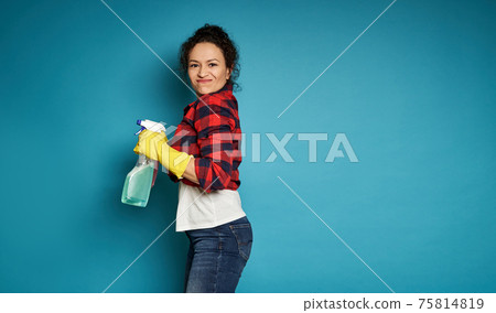 Side view to a mixed race woman holding a detergent cleaning spray and looking at the camera, posing over blue background with copy space 75814819