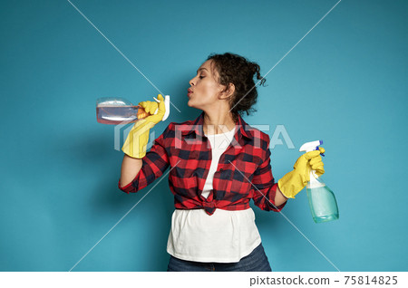 Attractive and playful young Latin American woman, a housewife, holding a cleaning spray in her hands like a gun and blowing off gunpowder as if after a shot Attractive and playful young Latin American woman, a housewife, holding a cleaning spray in her hands like a gun and blowing off gunpowder as if after a shot 75814825