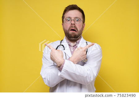 Bizarre, stunned male doctor points index fingers in different directions and looks with a strange Bizarre, stunned male doctor points index fingers in different directions and looks with a strange 75823490