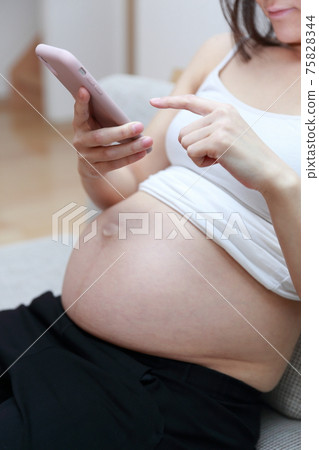 Pregnant woman sitting on the couch and operating a smartphone 75828344