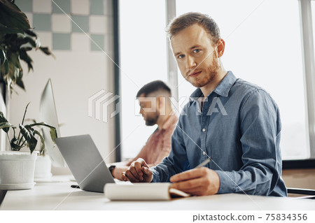 Young businessman making notes in notepad while sitting at the working table in modern office Young businessman making notes in notepad while sitting at the working table in modern office 75834356