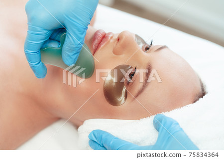 Beautiful woman having guasha scraper face massage done by cosmetologist Beautiful woman having guasha scraper face massage done by cosmetologist 75834364