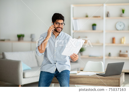 Home office. Confident arab entrepreneur talking with companion on phone and checking financial reports Home office. Confident arab entrepreneur talking with companion on phone and checking financial reports 75837163