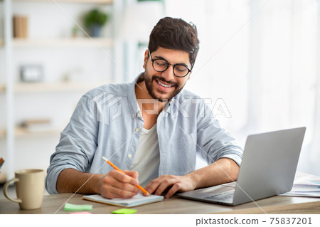 Happy arab man writing business report on laptop and taking notes, sitting at workplace at home interior Happy arab man writing business report on laptop and taking notes, sitting at workplace at home interior 75837201