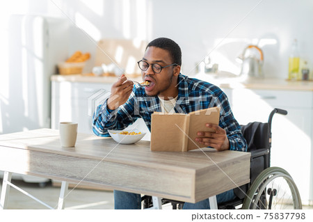 Young black disabled guy in wheelchair eating breakfast and reading book at kitchen. Stay home pastimes 75837798