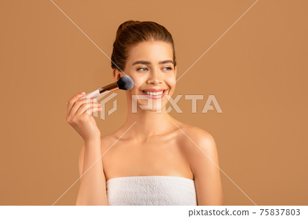Happy millennial woman with perfect skin applying blusher or powder, using makeup brush over brown studio background Happy millennial woman with perfect skin applying blusher or powder, using makeup brush over brown studio background 75837803