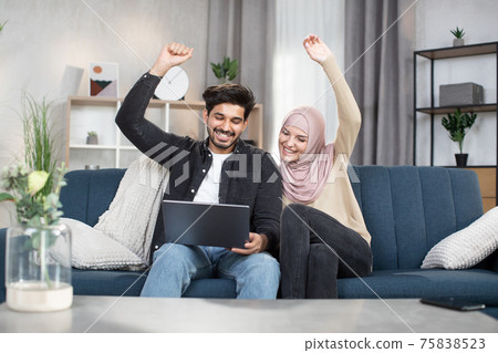 Cheerful young Muslim couple, holding hands up in victory gesture, celebrating success. Arabian couple using laptop for watching sports match together sitting on comfortable sofa in cozy living room Cheerful young Muslim couple, holding hands up in victory gesture, celebrating success. Arabian couple using laptop for watching sports match together sitting on comfortable sofa in cozy living room 75838523