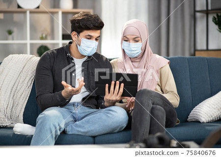 Handsome Arabian man in protective mask, talking and gesturing to his Muslim girlfriend in hijab and mask, sitting at home and discussing information they read from tablet. Covid-19 isolation concept Handsome Arabian man in protective mask, talking and gesturing to his Muslim girlfriend in hijab and mask, sitting at home and discussing information they read from tablet. Covid-19 isolation concept 75840676