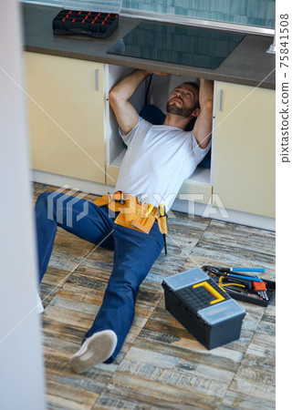 Stop leakage. Professional repairman, plumber wearing tool belt examining and fixing sink pipe in the kitchen indoors Stop leakage. Professional repairman, plumber wearing tool belt examining and fixing sink pipe in the kitchen indoors 75841508