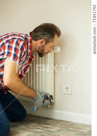 Quality help. Handyman using pipe wrench while repairing radiator battery in the room. Heating restoration 75841702