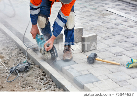 The bricklayer uses a grinder and diamond cutting discs to cut off the protruding part of the paving slabs for leveling on the sidewalk. 75844627