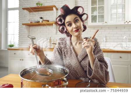 Young funny attractive housewife in curlers and bathrobe cooking at home kitchen Young funny attractive housewife in curlers and bathrobe cooking at home kitchen 75844939