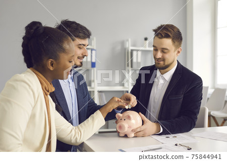 Happy couple together put money in piggy bank sitting at desk with banking advisor Happy couple together put money in piggy bank sitting at desk with banking advisor 75844941