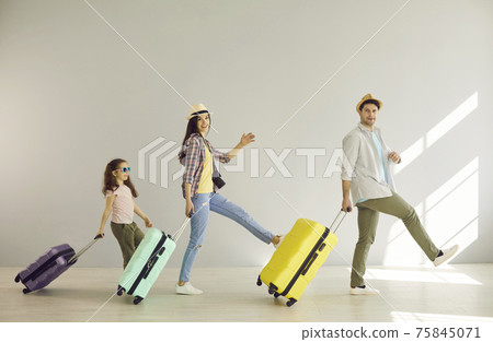 Cheerful mom, dad and their daughter have fun walking with suitcases along the gray wall. Cheerful mom, dad and their daughter have fun walking with suitcases along the gray wall. 75845071