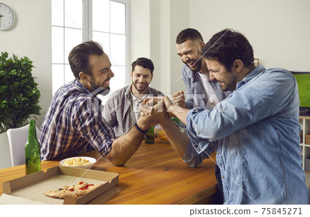Cheerful company of male friends at home drinking beer and competing in arm wrestling. Cheerful company of male friends at home drinking beer and competing in arm wrestling. 75845271