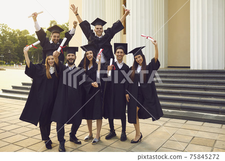 Cheerful university graduates posing with dimplomas in raised hands over university building 75845272