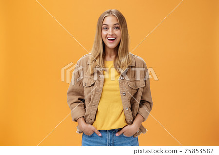 Charismatic tanned woman in corduroy jacket and yellow t-shirt ready for chilly autumn walk with friends smiling joyfully gazing entertained at camera holding hand in pockets casually over orange wall Charismatic tanned woman in corduroy jacket and yellow t-shirt ready for chilly autumn walk with friends smiling joyfully gazing entertained at camera holding hand in pockets casually over orange wall 75853582
