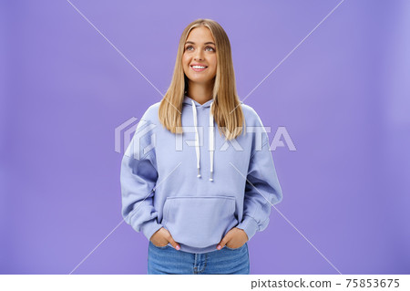 Winter comming. Dreamy kind and cute young girl with straight fair hair and tan wearing hoodie looking at upper left corner with nice white smile and admiring expression holding hand in pockets 75853675