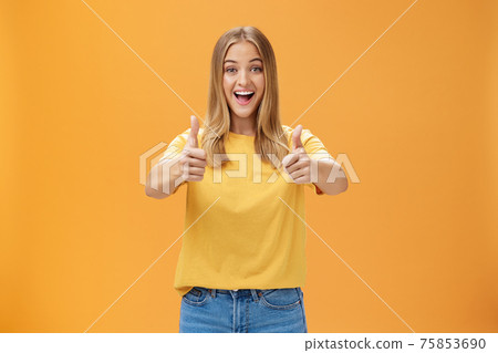 Woman supports with raised thumbs up and amused cheerful smile showing positive attitude expressing like on concept or idea giving approval posing happy and delighted against orange background 75853690