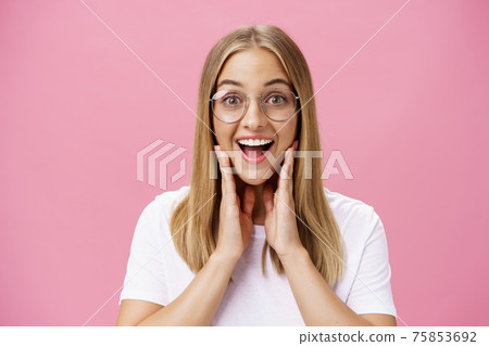 Woman liking new glasses picked in store feeling happy finding right frame touching cheeks from happiness smiling cheerfully at camera wearing transparent eyewear and t-shirt over pink wall 75853692