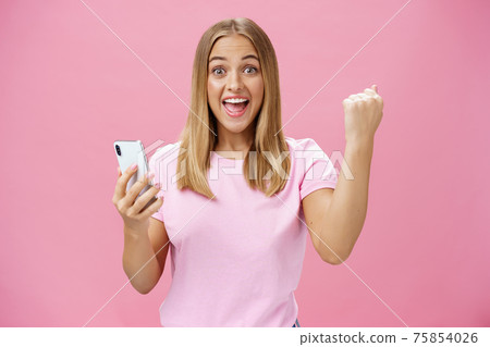 Girl beat own record in smartphone game raising clenched fist in cheer and triumph holding cellphone, smiling excited and happy at camera celebrating victory with joyful gesture over pink background 75854026