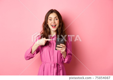 Young woman pointing finger at her phone and smiling, talking about smartphone app, showing something interesting online, standing over pink background Young woman pointing finger at her phone and smiling, talking about smartphone app, showing something interesting online, standing over pink background 75854087