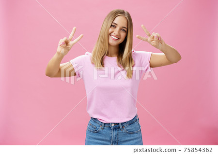 Cute chubby carefree young woman with short fair hair tilting head showing peace gestures with happy friendly smile having fun spending time amused and joyful against pink background 75854362